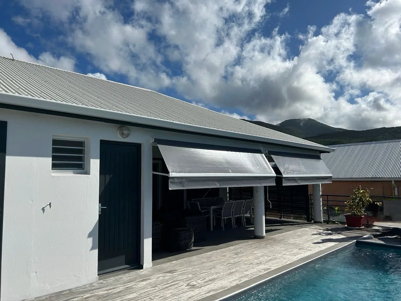 Villa avec deux stores bras droits gris au-dessus d'une terrasse et d'une piscine, montagne en fond